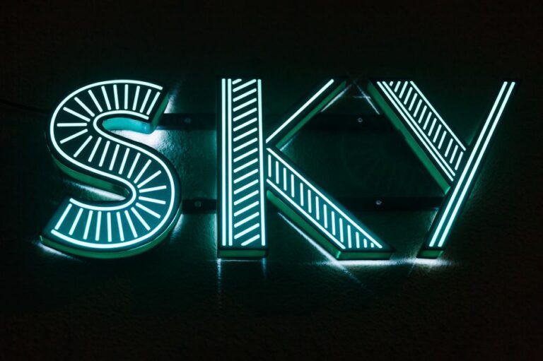 Illuminated building letters glowing at dusk beside a sidewalk, with sheltered wiring and evenly lit sign faces